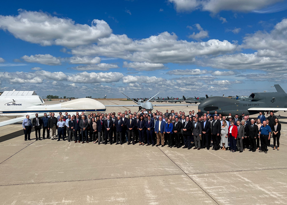 a group of around 100 people stand in front of black hawk drones at grand sky