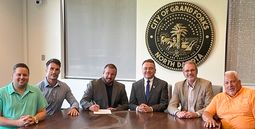 In front of the City of Grand Forks North Dakota seal at The HIVE in Downtown, sit, from left to right, Johnny Ryan, Mayor Brandon Bochenski, Mike Furmanski, Ned Tabat, Keith Lund, and Bill Adams. Ned Tabat holds a pen while signing a piece of paper that signifies the opening of Meadowlark Aircraft Company.