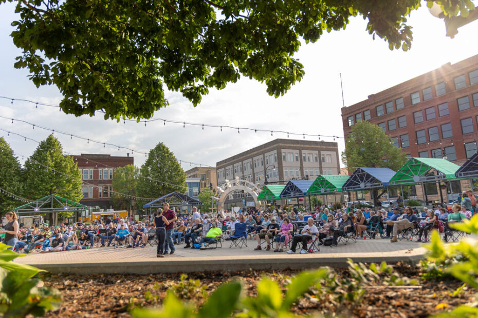 Grand Forks North Dakota town square is filled with a large crowd of people sitting in lawn chairs and dancing at the 2023 Blues on the Red music festival while the sun sets over the city.