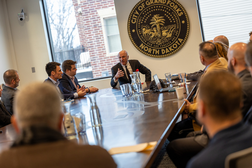 Kevin O'Leary sits at the head of the table in The HIVE with the City of Grand Forks seal behind him.